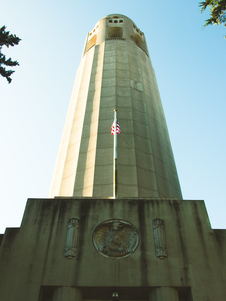Looking straight up at Coit Tower in San Francisco, an American flag on a pole against the fluted concrete column and a clear blue sky