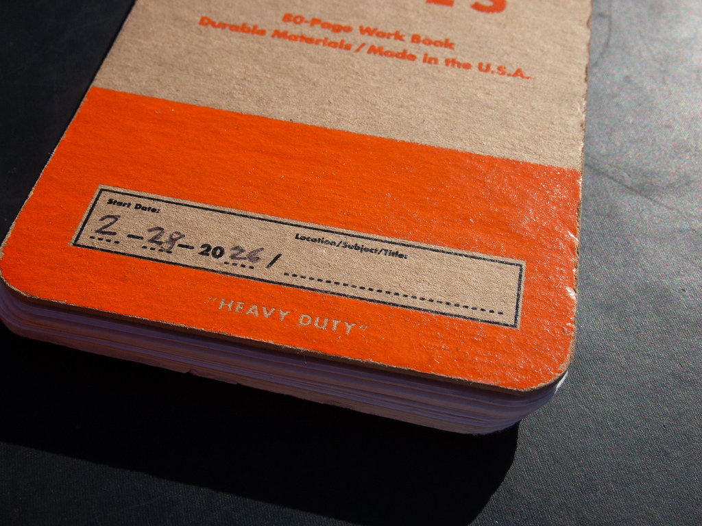 Close-up of a pocket notebook cover in kraft and bright orange, the start date 2-29-2026 written by hand, in hard sunlight on a dark surface