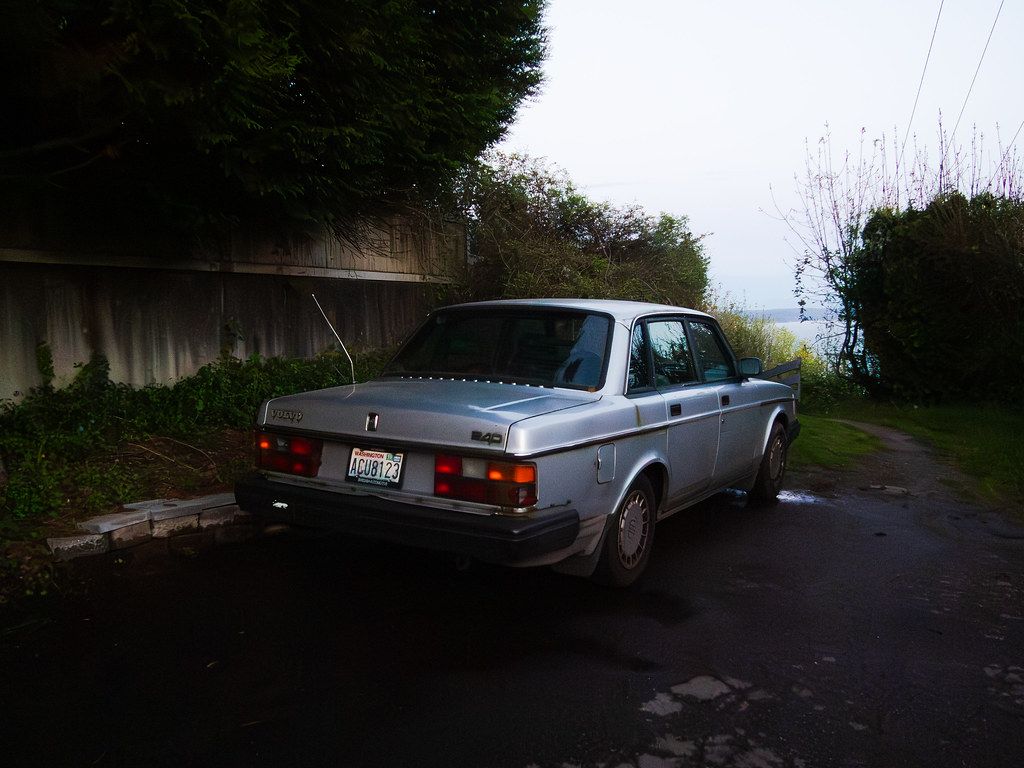 A silver Volvo 240 sedan parked at dusk at the end of a wet lane, framed by dark hedges and a weathered fence, with gray water in the distance